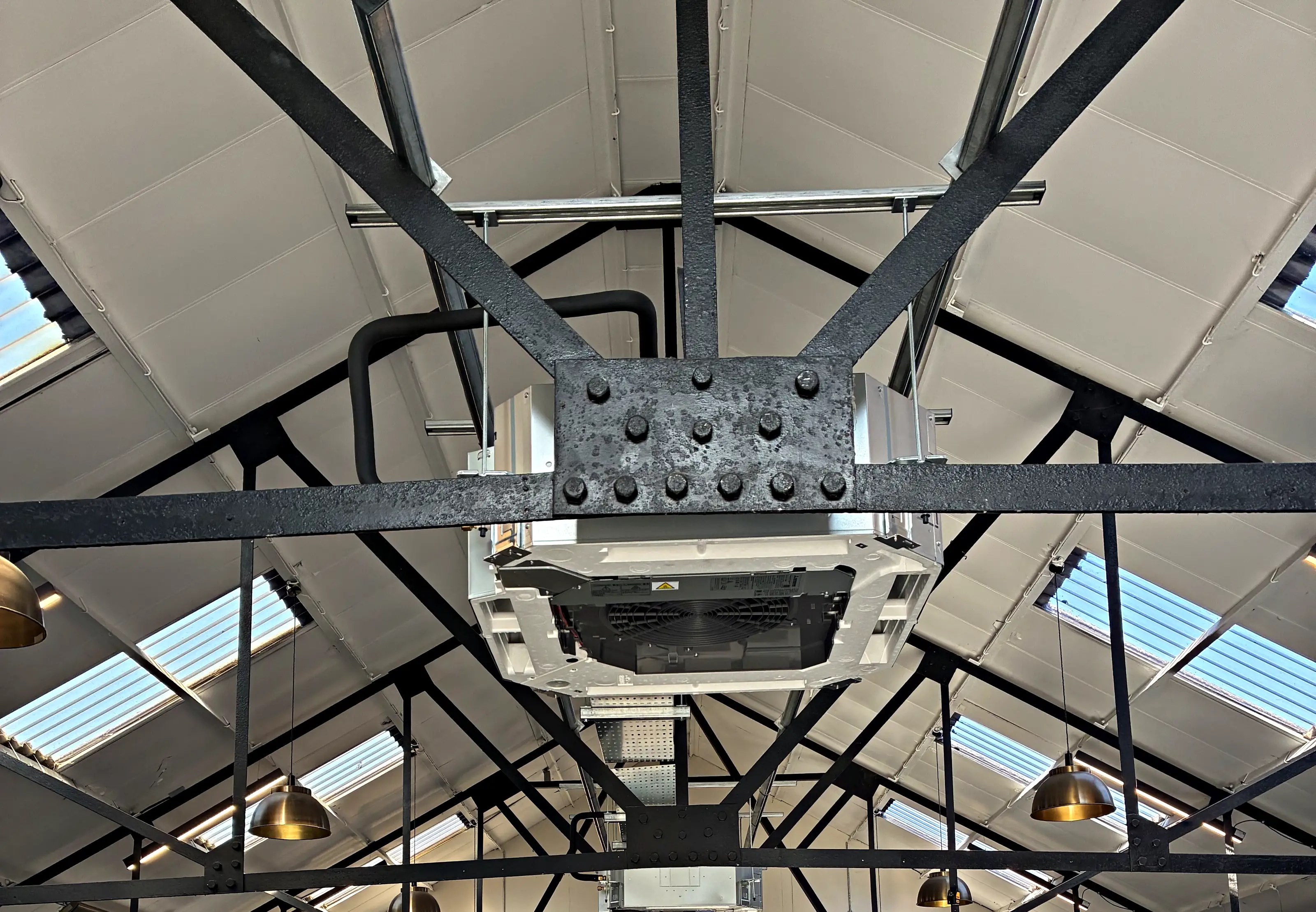 A ceiling cassette hung beneath a bolted steel roof truss, with its pipework run along the beam