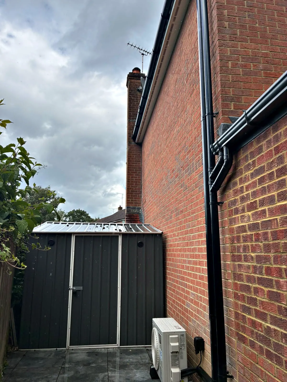 An outdoor unit in the side return of a brick house, its pipework running up the wall beside the downpipe